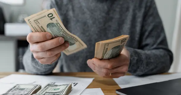 Person counting money with hands, holding stacks of ten-dollar bills on a table with documents